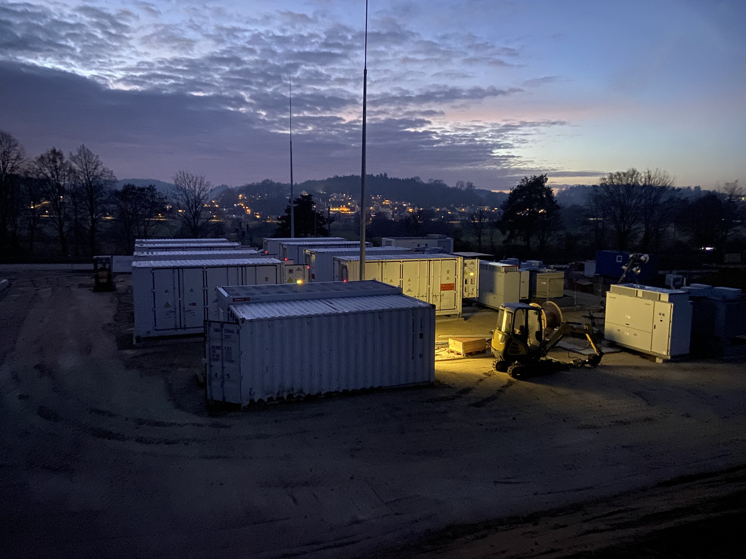 Batteriespeicherpark in der Abenddämmerung: Speichercontainer auf der Baufläche, ein beleuchteter Minibagger, im Hintergrund Hügel mit den Lichtern eines Ortes