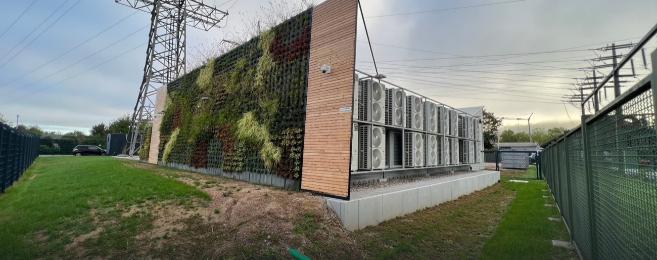Rechenzentrumsgebäude mit begrünter Fassade, Holzverkleidung und Reihen von Klimageräten, daneben ein Strommast